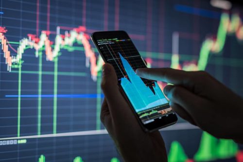 Bolsa de Valores: em primeiro plano uma pessoa acompanhando as oscilações do mercado financeiro na tela do celular e em segundo plano está mostrando o gráfico de oscilações da bolsa de valores.