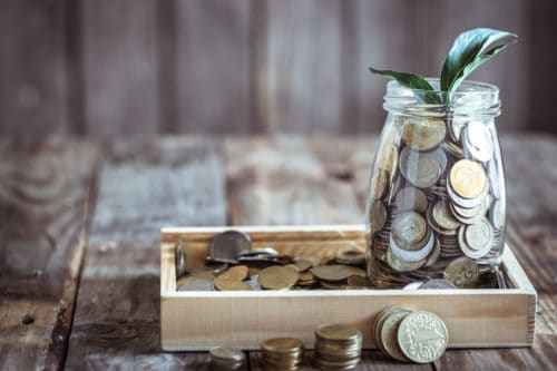 fundos de investimento: uma caixa de madeira cheia de moedas e um pote de vidro com várias moedas dentro e uma planta.