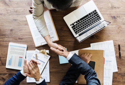 investimento anjo: duas pessoas realizando um acordo com um aperto de mão. Um notebook e algumas folhas de papéis sob a mesa.