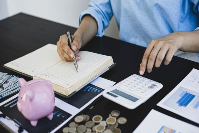 carteira de dividendos: um homem fazendo anotações no caderno e fazendo contas usando a calculadora. Na mesa tem várias moedas e planilhas.