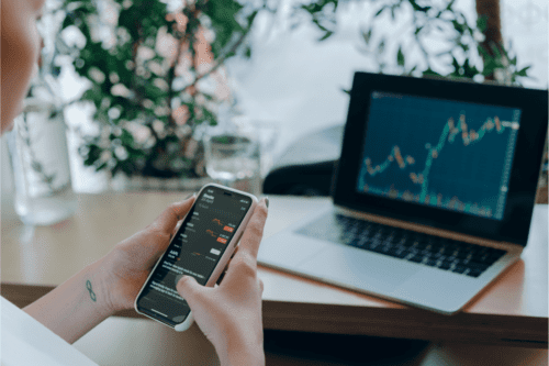 como começar a investir: uma pessoa acompanhando a oscilação do mercado financeiro no celular e no notebook.