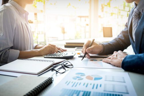 derivativos financeiros: uma mesa cheia de papel com gráficos e caderno, uma calculadora e duas pessoas fazendo anotações.