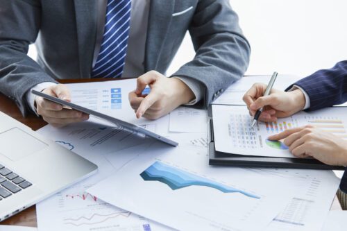 Pessoas de terno escrevem e apontam para papéis com gráficos e números na mesa para ilustrar a construção de um plano de investimento