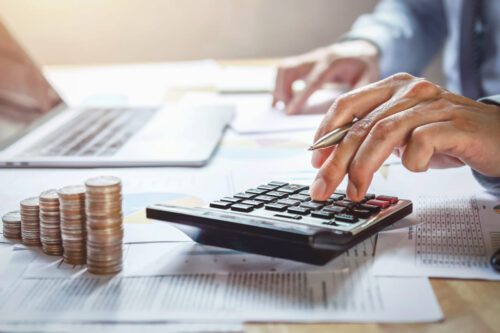 o que é ipca: uma pessoa digitando em uma calculadora e uma pilha de moedas ao lado, mais um notebook.