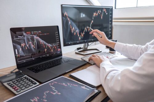 Opções binárias: pessoa adulta branca, blusa social branca, aponta para tela de computador com gráfico em mesa com dois monitores, calculadora e papéis