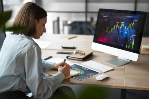 trader: uma mulher escrevendo na agenda em um escritório, sob a mesa um computador e algumas folhas de papéis e canetas.