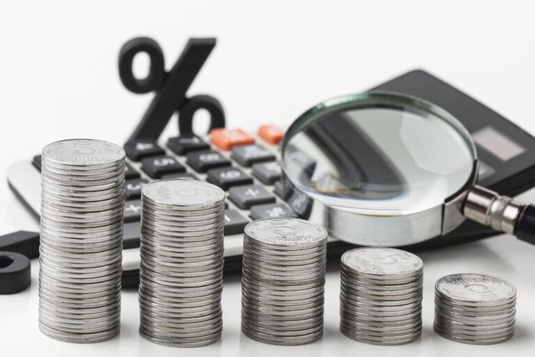 taxa de juros real: pilha de moedas na cor prata com uma lupa, uma calculadora em segundo plano.