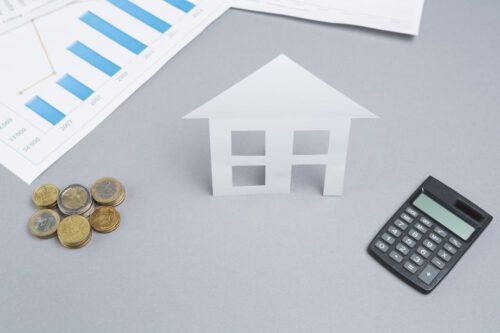 Fundos imobiliários híbridos: casa de papel recortada sob lugar cinza ao lado de papel branco com gráfico, moedas e calculadora