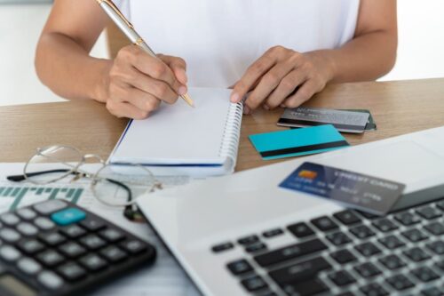 IOF: uma mulher fazendo anotações com uma calculadora e um notebook ao lado mais alguns cartões de crédito.