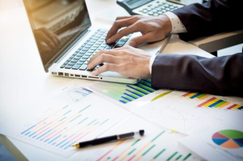 análise fundamentalista: uma pessoa digitando no notebook e gráficos e folhas em cima da mesa e uma caneta e calculadora.