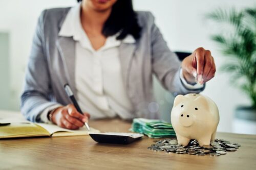 como economizar dinheiro: uma mulher colocando moedas no cofrinho e fazendo anotações.
