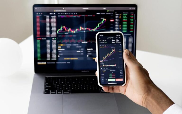 Mão segurando celular exibindo gráfico dos índices financeiros na tela. Em segundo plano, um notebook exibindo gráficos em sua tela.