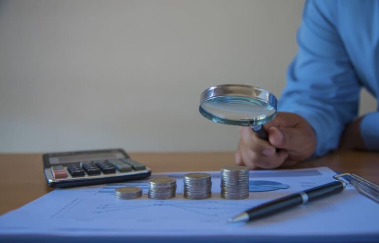Mão de homem segurando lupa apontando para papel que tem calculadora, moedas empilhadas e canetas por cima.