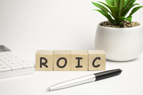 Cubos de madeira com letras formando a sigla ROIC sobre uma mesa com caneta, calculadora e vaso de planta.