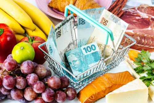 Uma esta de mercado com notas de dinheiro dentro, envolta da cesta encontram-se frutas e proteinas