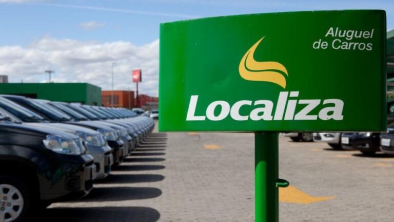 Placa verde com logo da Localiza em branco e amarelo na frente de m estacionamento cheio de carros