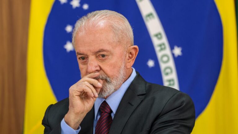 atual presidente Lula de terno com a mão na boca na frente de uma bandeira do Brasil