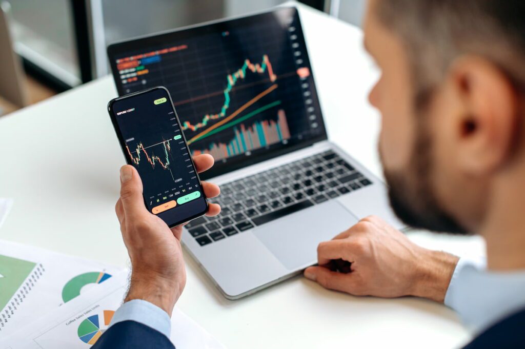 Homem com notebook e celular analisando gráficos das melhores empresas para investir em 2024.