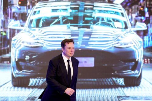 Elon Musk de terno e gravata na frente de uma tela com uma imagem de um carro