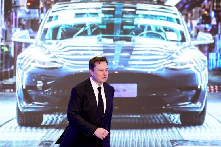 Elon Musk de terno e gravata na frente de uma tela com uma imagem de um carro