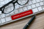 Óculos e caneta sobre mesa de madeira ao lado de teclado com etiqueta vermelha MERGERS & ACQUISITIONS (M&A).