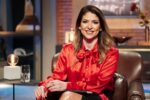 Carol Paiffer, vestida com uma blusa vermelha brilhante e sentada em uma poltrona de couro, sorrindo em um ambiente elegante.