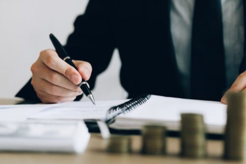 Pessoa de terno escrevendo em um caderno com moedas ao lado, simbolizando preparação para a certificação CEA.