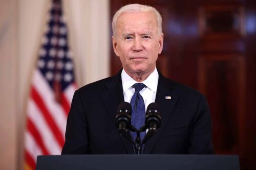 Joe Biden fazendo um discurso, com uma bandeira dos EUA desfocada ao fundo, usando terno escuro e gravata.