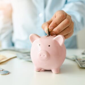 Pessoa depositando uma moeda em um cofrinho rosa com notas e moedas ao fundo, representando o Tesouro Selic 2027 ou 2029.