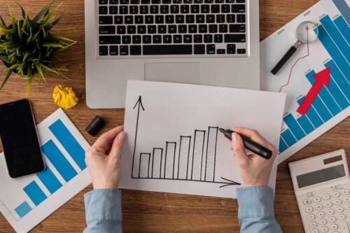 Pessoa desenhando gráfico de crescimento ao lado de laptop e planilhas, representando metas financeiras e objetivos financeiros.