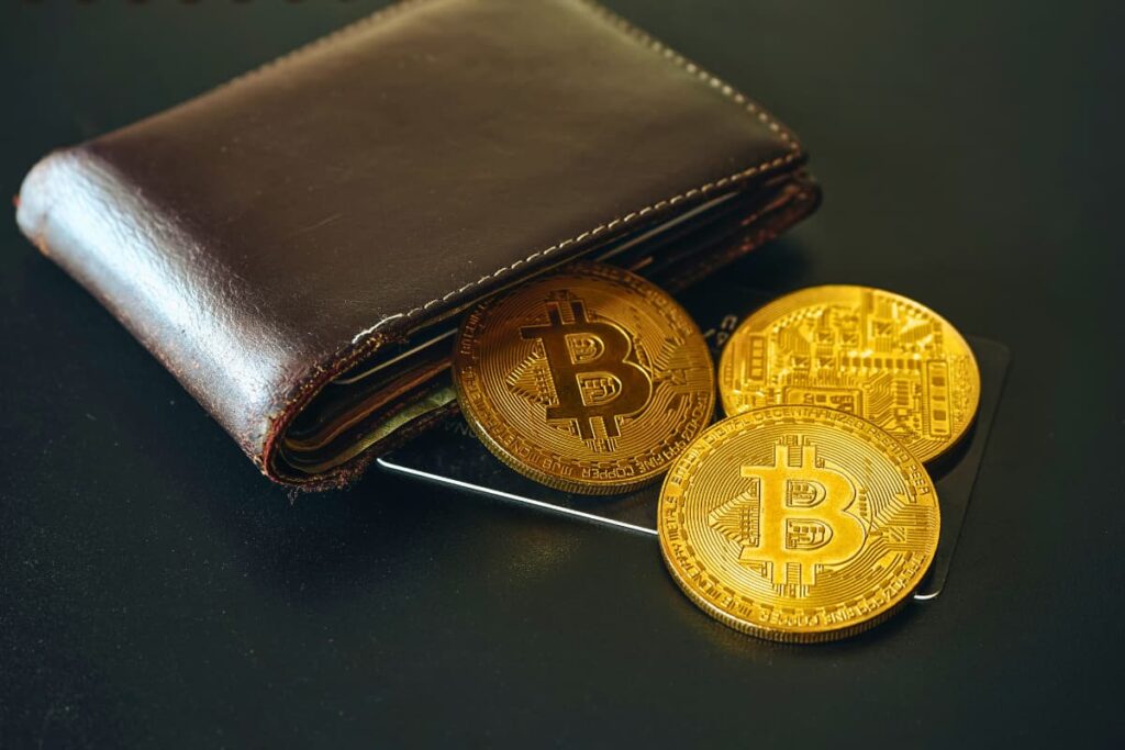 Imagem de uma carteira Bitcoin de couro com três moedas físicas de Bitcoin e um cartão, representando o armazenamento de criptomoedas.
