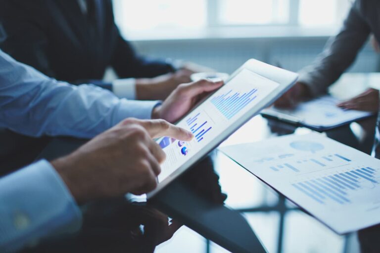 Profissionais analisam gráficos em um tablet, discutindo estratégias de investimento, capex e opex em uma reunião corporativa.