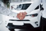 Aperto de mãos entre duas pessoas em frente a um carro branco, simbolizando um acordo e como vender um carro financiado.