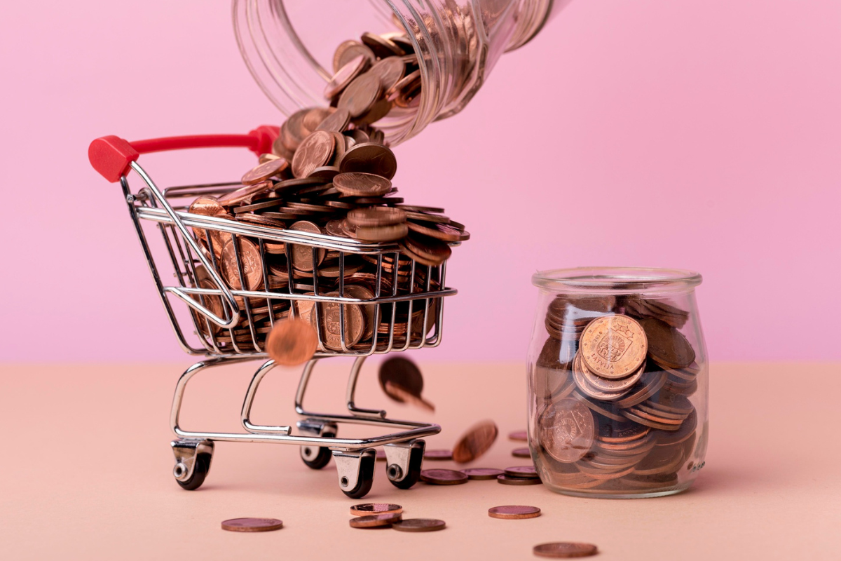 m minicarrinho de supermercado de metal sendo preenchido por moedas douradas que caem de um pote de vidro virado.