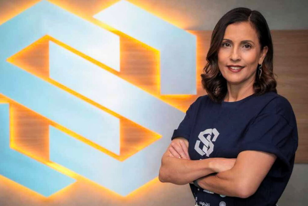 Ana Karina Bortoni, Presidente do Grupo Silvio Santos, de braços cruzados, sorrindo, com logo iluminado ao fundo.