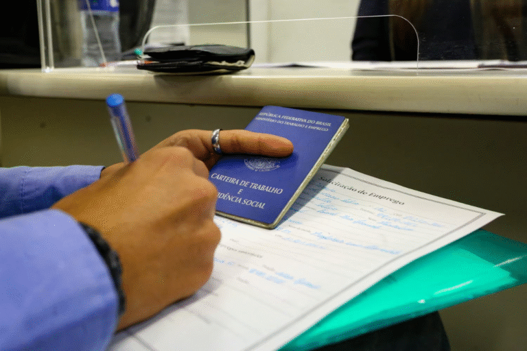 Pessoa segurando uma carteira de trabalho e uma caneta, com documentos na mesa, representando vagas de emprego oferecidas pela Prefeitura de São Paulo.