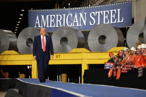 Imagem de Donald Trump em uma fábrica de aço, com rolos de aço ao fundo e trabalhadores usando capacetes e roupas de segurança.
