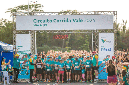 Participantes na Corrida Vale 2024 em Vitória, ES, reunidos na largada da corrida com inscrições de corrida visíveis e um grande banner acima com o nome do evento.