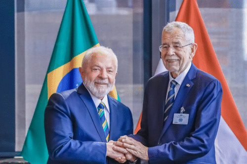 Duas pessoas em traje formal apertando as mãos em frente às bandeiras do Brasil e de outro país, simbolizando acordo ou cooperação internacional.