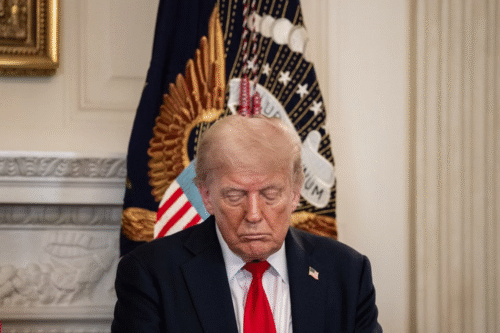 Imagem de Donald Trump com expressão séria, usando terno escuro e gravata vermelha, em um ambiente formal com bandeira e decoração ao fundo.