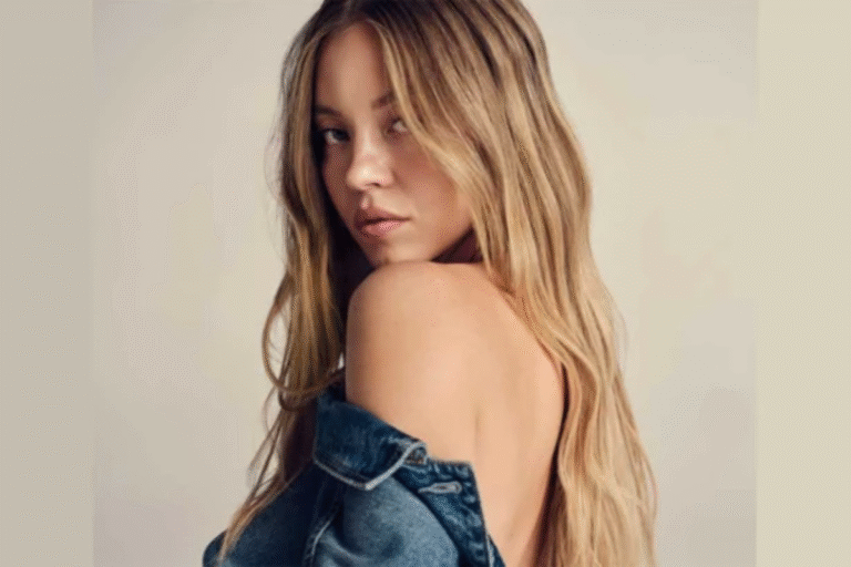 Retrato de Sydney Sweeney com cabelo loiro e expressão sedutora, usando jaqueta jeans caída no ombro, em fundo neutro, destacando beleza e estilo feminino.