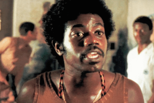 Imagem de um homem negro com cabelo afro e expressão séria, em ambiente com outras pessoas ao fundo, vestindo camiseta marrom e colar de contas, retrato de um personagem ou pessoa famosa.