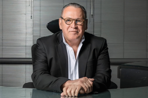 Sidney Oliveira de negócios sério sentado na mesa de escritório, usando terno preto e óculos, com expressãoconfiante.