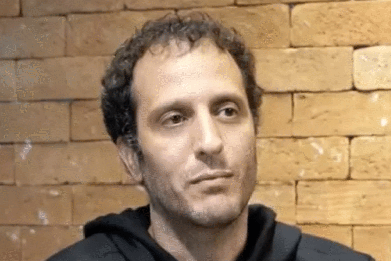 Imagem de um homem com cabelo cacheado e expressão pensativa, vestindo uma blusa preta, contra um fundo de tijolos. Perfeito para conteúdo de moda, beleza ou estilo de vida masculino.