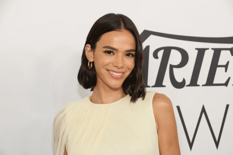Bruna Marquezine sorridente com cabelo escuro, usando blusa branca, em evento com fundo branco e logotipo, promovendo beleza e autoestima.