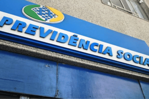 Fachada de agência da Previdência Social ao ar livre, com placa azul com letras em relevo destacando 'Previdência Social'. Edifício de altura média ao fundo.
