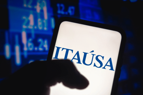 Imagem de um smartphone sendo segurado, mostrando a palavra 'ITAÚSA' na tela, com fundo de telas com gráficos financeiros. Ideal para conteúdos de investimentos, ações e mercado financeiro.