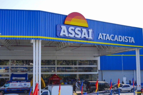 Fachada da loja Assaí Atacadista com fachada azul, logo colorido e ampla entrada, com pessoas e veículos na área de estacionamento.