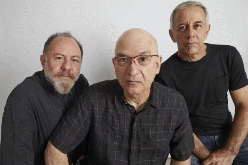 Três homens adultos posando juntos em uma foto de estúdio, roupa casual e expressão séria, com foco na amizade e na presença de pessoas maduras.