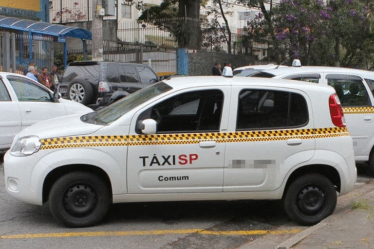 Táxi comum de São Paulo estacionado na rua, com outros veículos ao fundo. Imagem mostra foto de táxi branco com detalhes em amarelo e vermelho, com foco em transporte urbano.